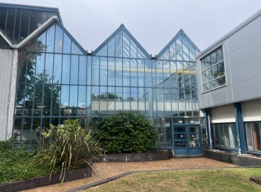 Glass-walled building with triangular roof peaks and blue framing; brick planters and greenery in the foreground.
