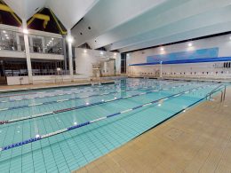 Indoor swimming pool with multiple lane ropes, starting blocks, and a viewing gallery on the left; bright ceiling lights and large windows.