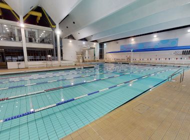 Indoor swimming pool with multiple lane ropes, starting blocks, and a viewing gallery on the left; bright ceiling lights and large windows.