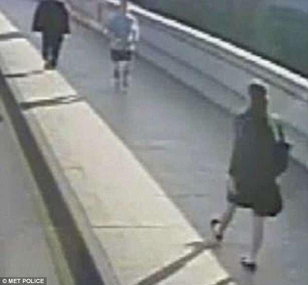 CCTV image of a curved pedestrian walkway with two people walking apart: a person in a dark coat and backpack on the right, and another person in a light-colored top and shorts toward the center, with a railing along the left side.