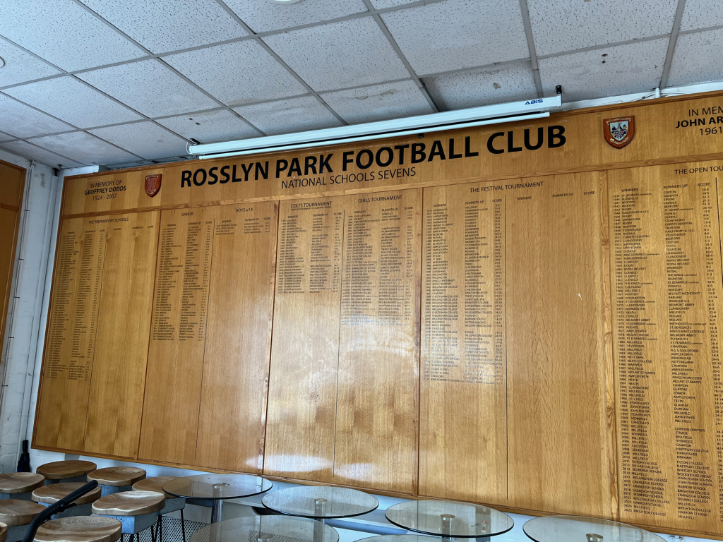 Wooden wall of name plaques at Rosslyn Park Football Club, with a large header and many names listed above seating area.