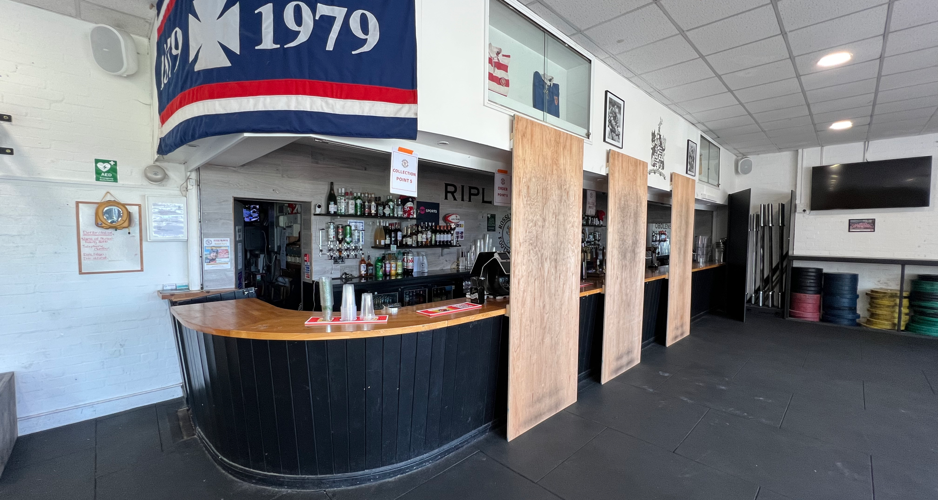 Bar interior with a curved black-and-wood counter, shelves of bottles behind, and tall wooden dividers; a large blue flag with red/white stripes and the number 1979 hangs from the ceiling above the bar.
