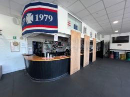 Bar interior with a curved black-and-wood counter, shelves of bottles behind, and tall wooden dividers; a large blue flag with red/white stripes and the number 1979 hangs from the ceiling above the bar.