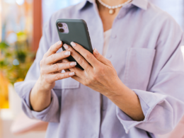 Person wearing a light blue shirt and a necklace holds a smartphone, looking at the screen indoors.