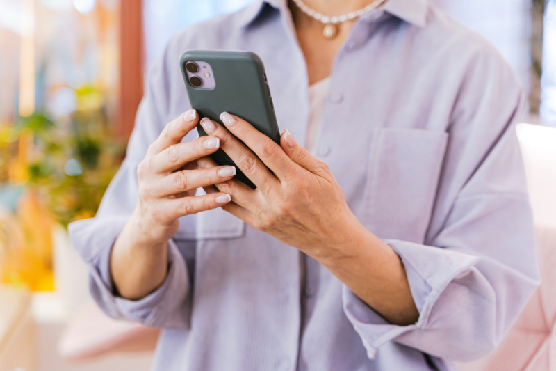 Person wearing a light blue shirt and a necklace holds a smartphone, looking at the screen indoors.