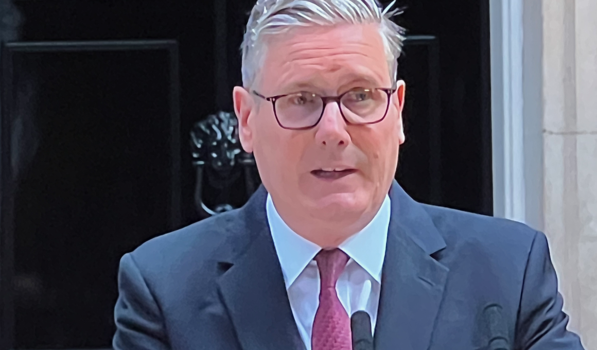 Middle-aged man in a navy suit and red tie, wearing glasses, speaking into a lapel microphone.