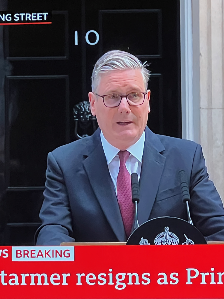 Man in a suit and glasses speaks at a podium with a red breaking news banner visible in the foreground