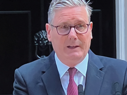 Middle-aged man in a navy suit and red tie, wearing glasses, speaking into a lapel microphone.