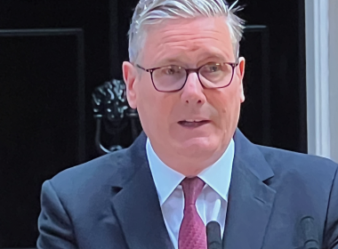 Middle-aged man in a navy suit and red tie, wearing glasses, speaking into a lapel microphone.