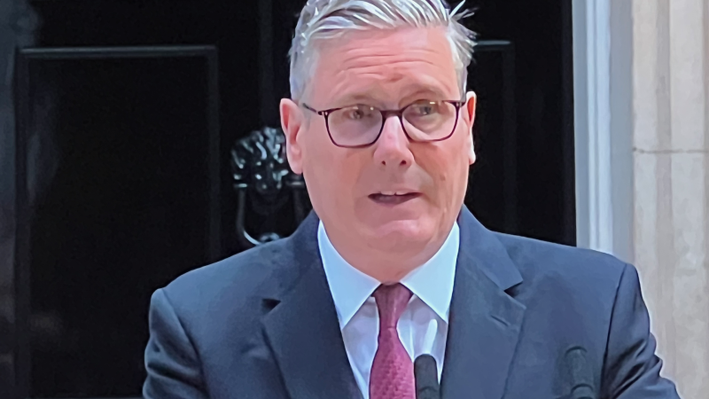 Middle-aged man in a navy suit and red tie, wearing glasses, speaking into a lapel microphone.