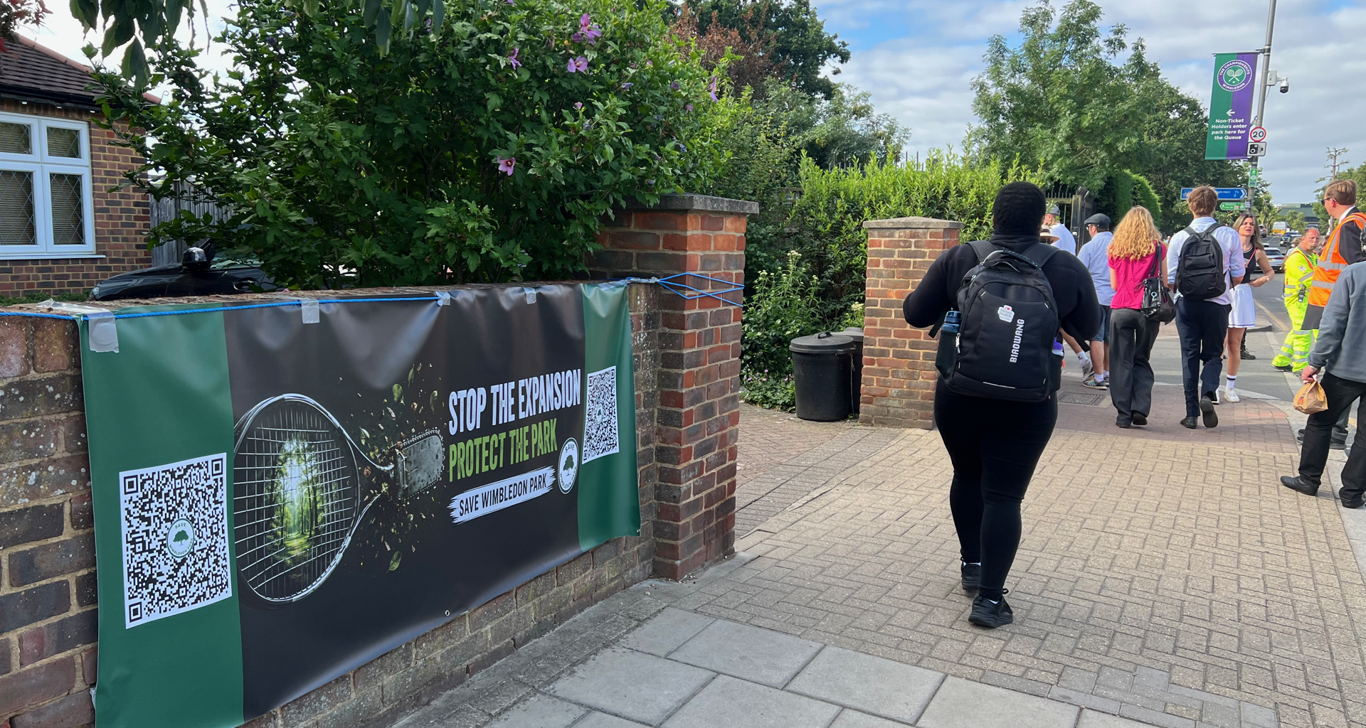 Banner on a brick wall reading 'Stop the expansion, protect the park' with QR codes, greenery, and pedestrians walking along a sidewalk on a sunny day.