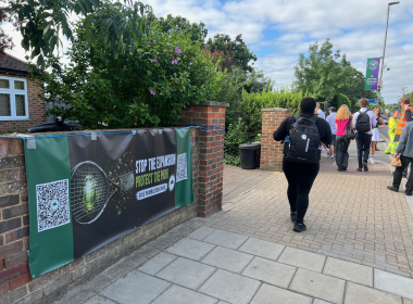 Banner on a brick wall reading 'Stop the expansion, protect the park' with QR codes, greenery, and pedestrians walking along a sidewalk on a sunny day.