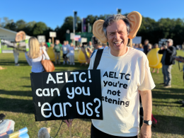 Simon Wright wearing large ear props, smiling, holding signs that say ‘AELTC! hear us?’ with others in background in a park.