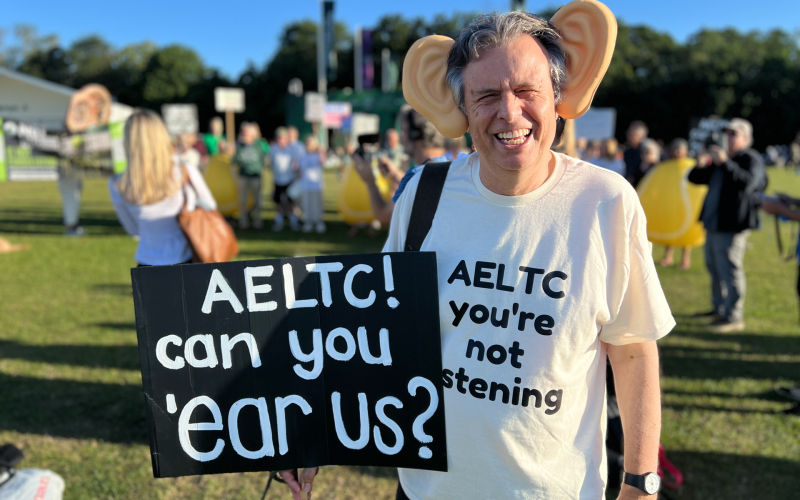 Simon Wright wearing large ear props, smiling, holding signs that say ‘AELTC! hear us?’ with others in background in a park.