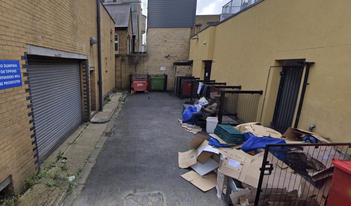 Narrow alley with brick buildings, overflowing cardboard boxes and dumpsters, and a man seated on a bench with belongings.