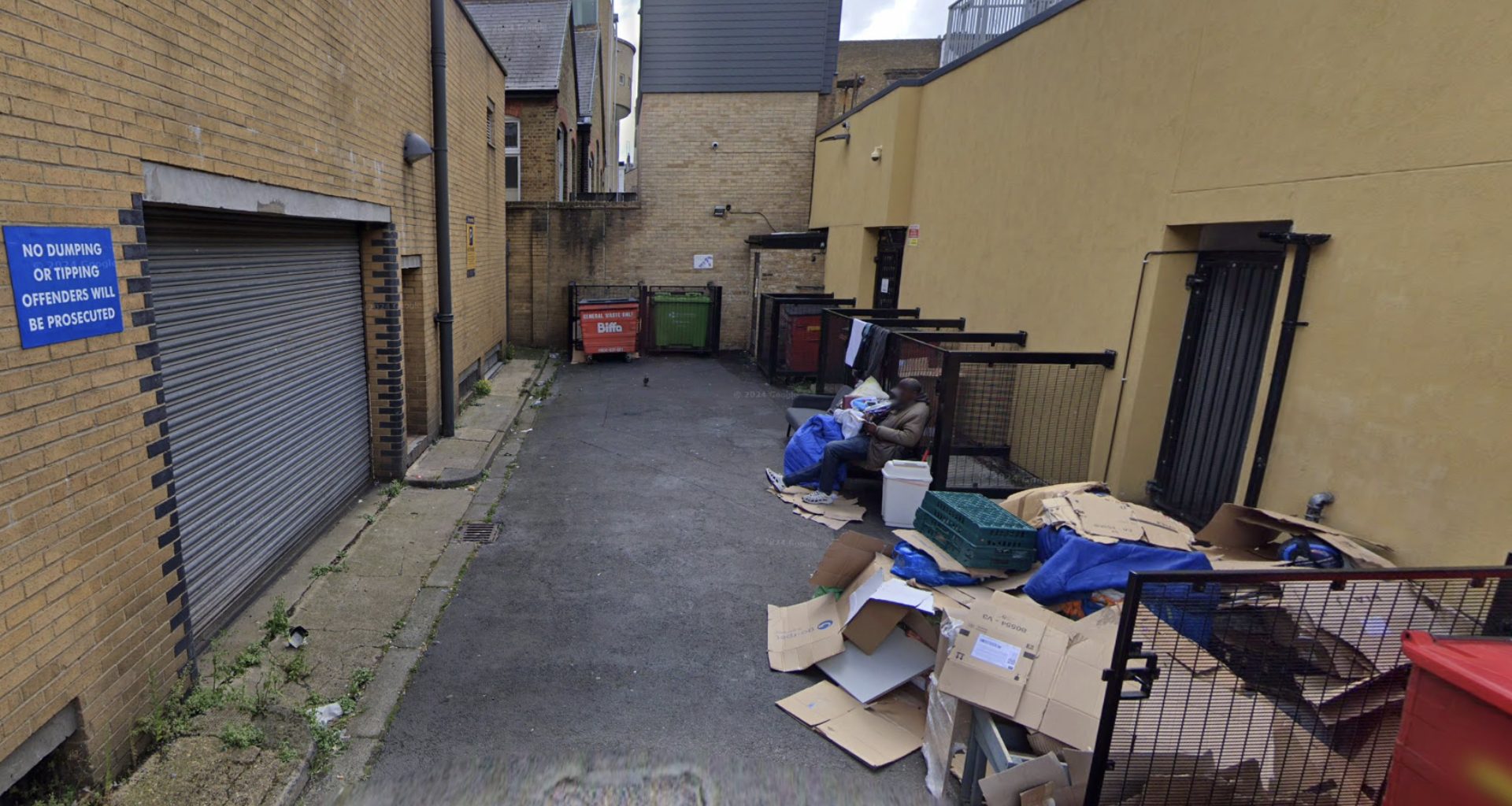 Narrow alley with brick buildings, overflowing cardboard boxes and dumpsters, and a man seated on a bench with belongings.