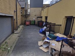 Narrow alley with brick buildings, overflowing cardboard boxes and dumpsters, and a man seated on a bench with belongings.