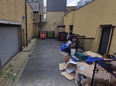 Narrow alley with brick buildings, overflowing cardboard boxes and dumpsters, and a man seated on a bench with belongings.