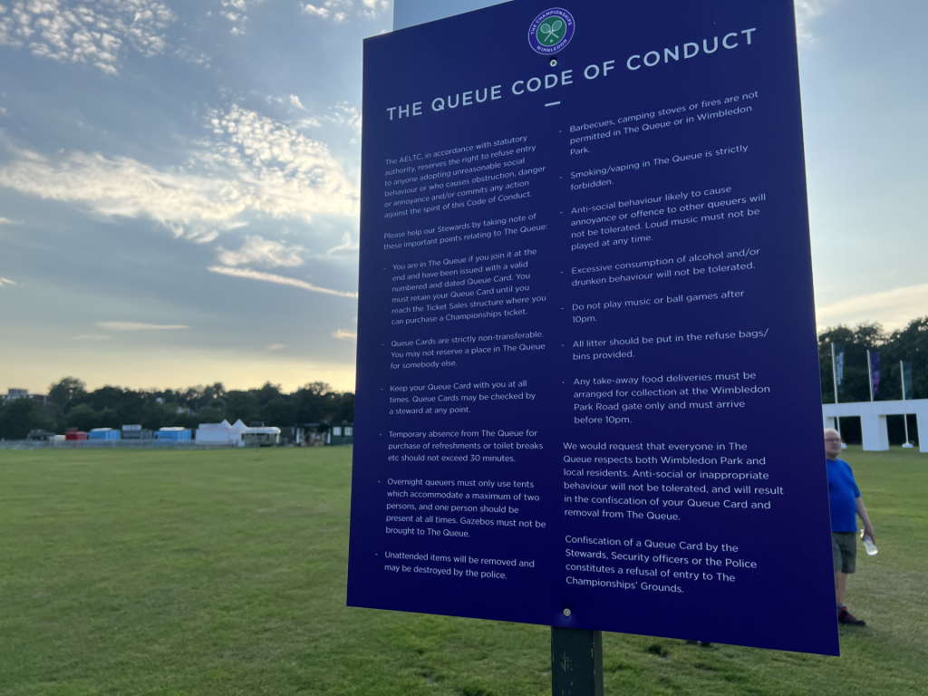 Large blue information board titled 'The Queue Code of Conduct' with Wimbledon logo, set in a grassy park with tents in the background.