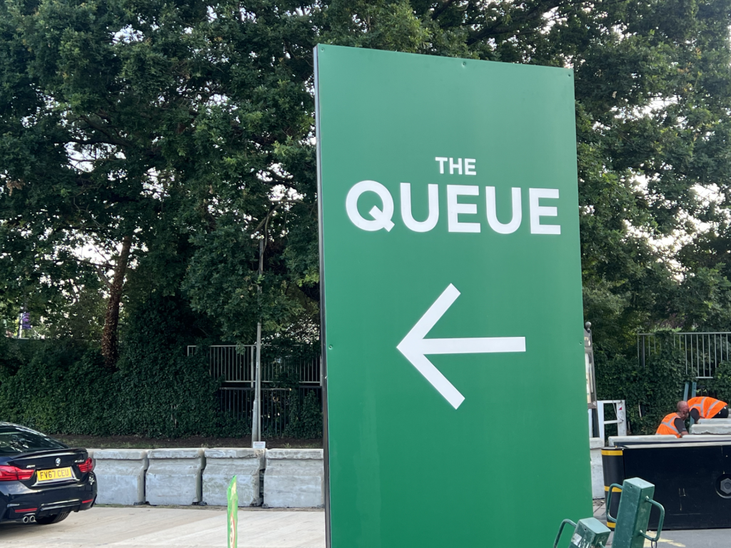 Green sign reading THE QUEUE with a left-pointing arrow, outdoors near a construction area.