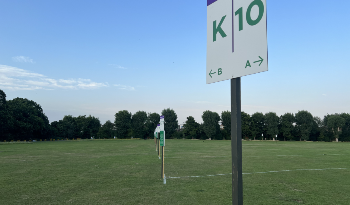 Tall sign on a pole in a grassy field reading 'Queue Section' with large 'K' and '10' and arrows 'A' and 'B' for directions below left and right.