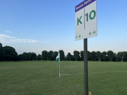 Tall sign on a pole in a grassy field reading 'Queue Section' with large 'K' and '10' and arrows 'A' and 'B' for directions below left and right.