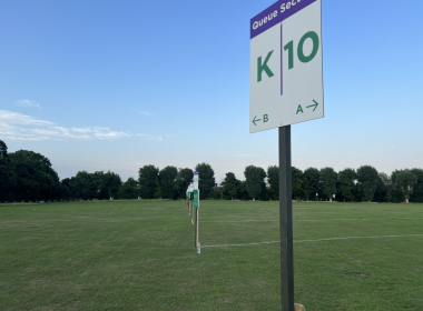 Tall sign on a pole in a grassy field reading 'Queue Section' with large 'K' and '10' and arrows 'A' and 'B' for directions below left and right.