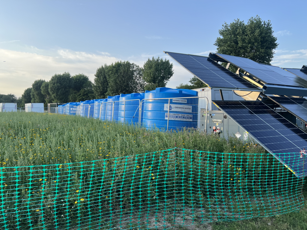 Solar panels on a tilted rack beside blue portable water tanks, fenced off in a grassy field with trees in the background.