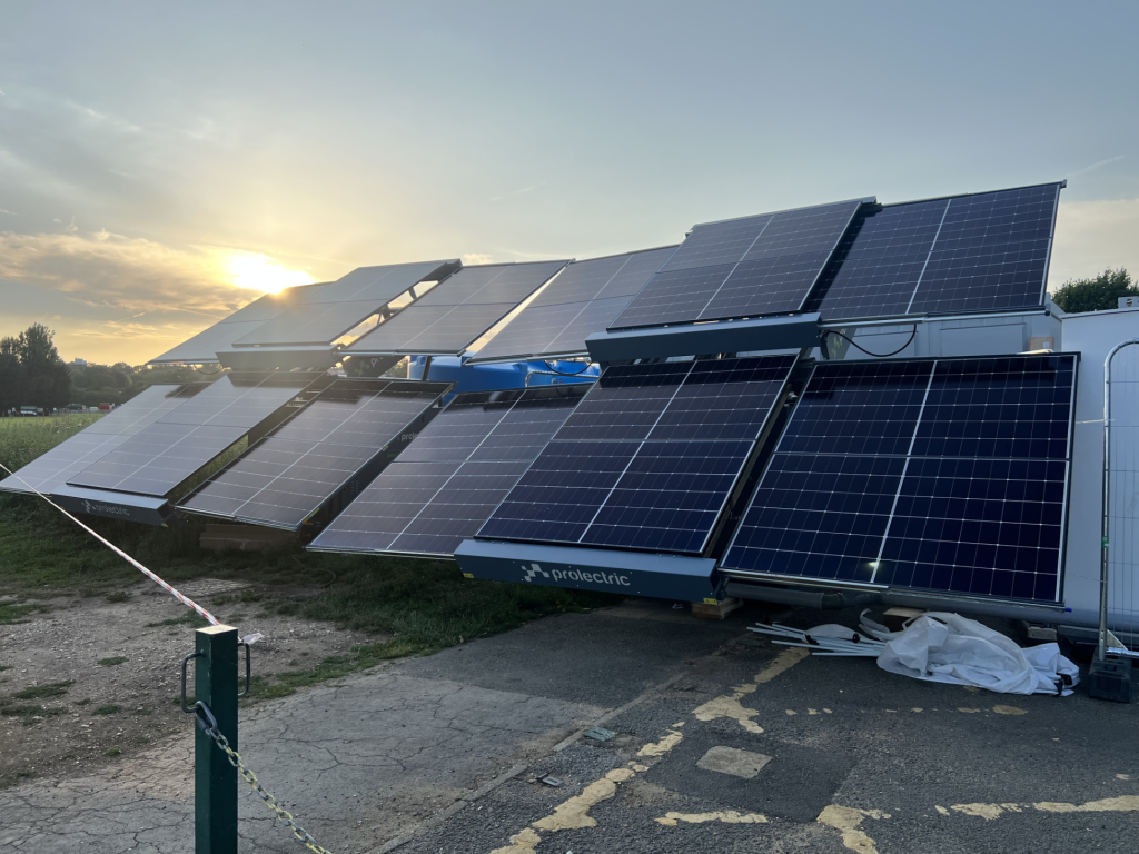 Row of solar panels tilted on metal frames outdoors at sunset, with a fence and construction debris nearby.