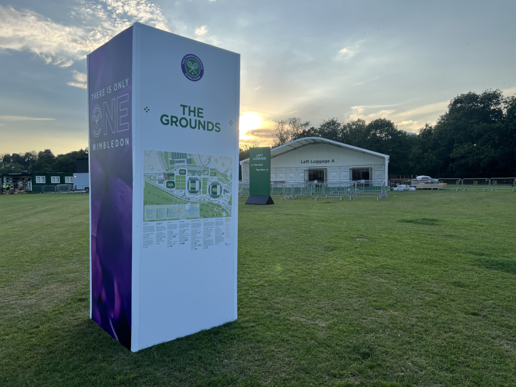 Large signboard with Wimbledon grounds map and 'THE GROUNDS' text at an outdoor event, sunset in the background.