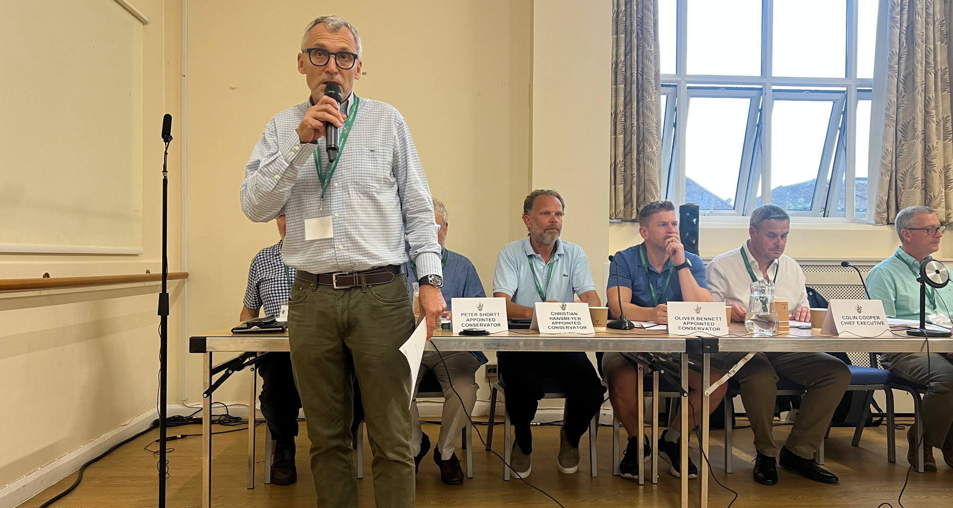 Man in a checkered shirt and glasses speaks into a handheld microphone at a panel discussion, with seated panelists and name placards behind him on a long table.