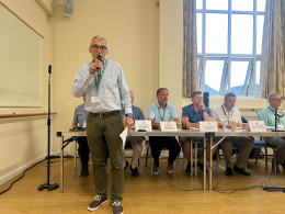 Man in a checkered shirt and glasses speaks into a handheld microphone at a panel discussion, with seated panelists and name placards behind him on a long table.