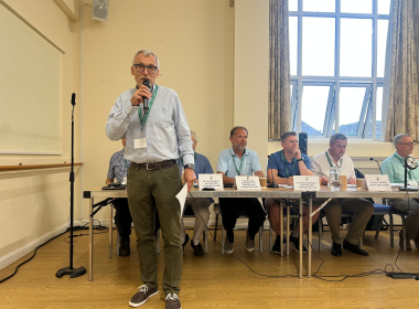 Man in a checkered shirt and glasses speaks into a handheld microphone at a panel discussion, with seated panelists and name placards behind him on a long table.