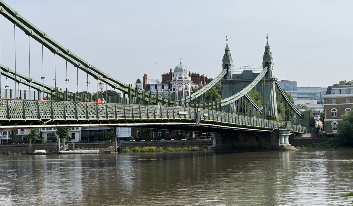 Hammersmith Bridge suspension bridge spans a river with lattice railings and decorative towers, city buildings visible in the background.