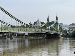 Hammersmith Bridge suspension bridge spans a river with lattice railings and decorative towers, city buildings visible in the background.