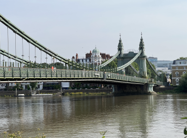 Hammersmith Bridge suspension bridge spans a river with lattice railings and decorative towers, city buildings visible in the background.