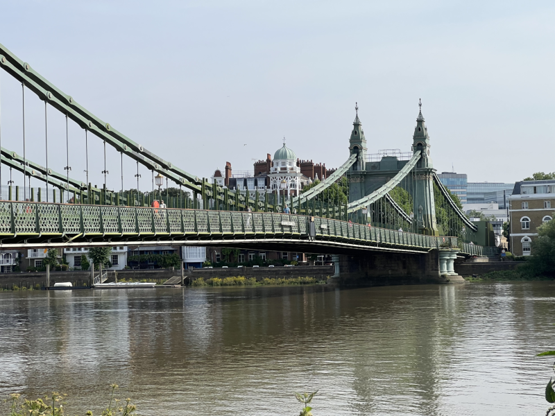 Hammersmith Bridge suspension bridge spans a river with lattice railings and decorative towers, city buildings visible in the background.