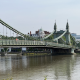 Hammersmith Bridge suspension bridge spans a river with lattice railings and decorative towers, city buildings visible in the background.