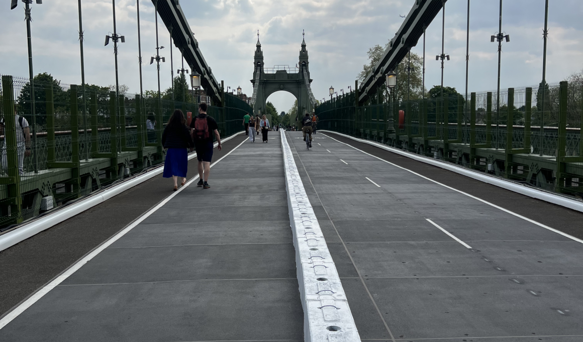 Pedestrian and cyclist walkway on a wide bridge, with a central white barrier rail and green side fencing, leading to ornate towers in the distance under a cloudy sky.