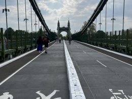 Pedestrian and cyclist walkway on a wide bridge, with a central white barrier rail and green side fencing, leading to ornate towers in the distance under a cloudy sky.