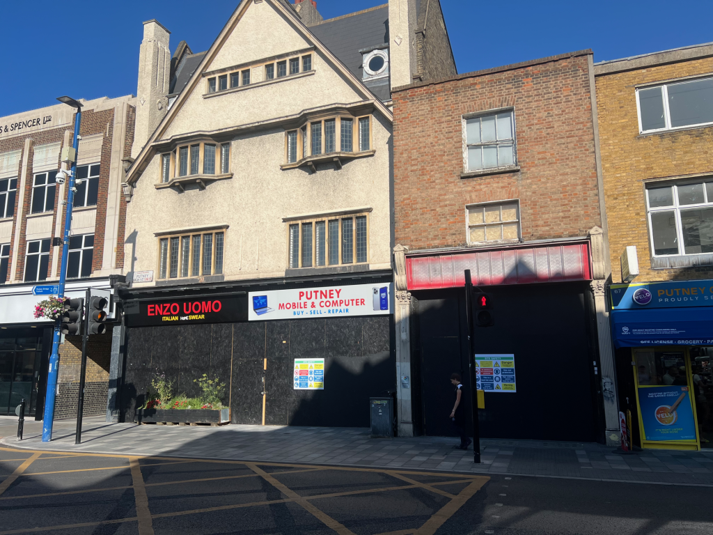 Shopfronts along a city street featuring ENZO UOMO and Putney Mobile & Computer signs, with an ornate beige building and blue sky above.