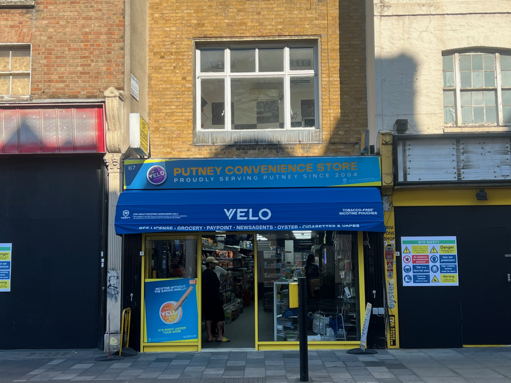 Putney Convenience Store storefront with a blue VELO sign and yellow entrance, customers inside visible.