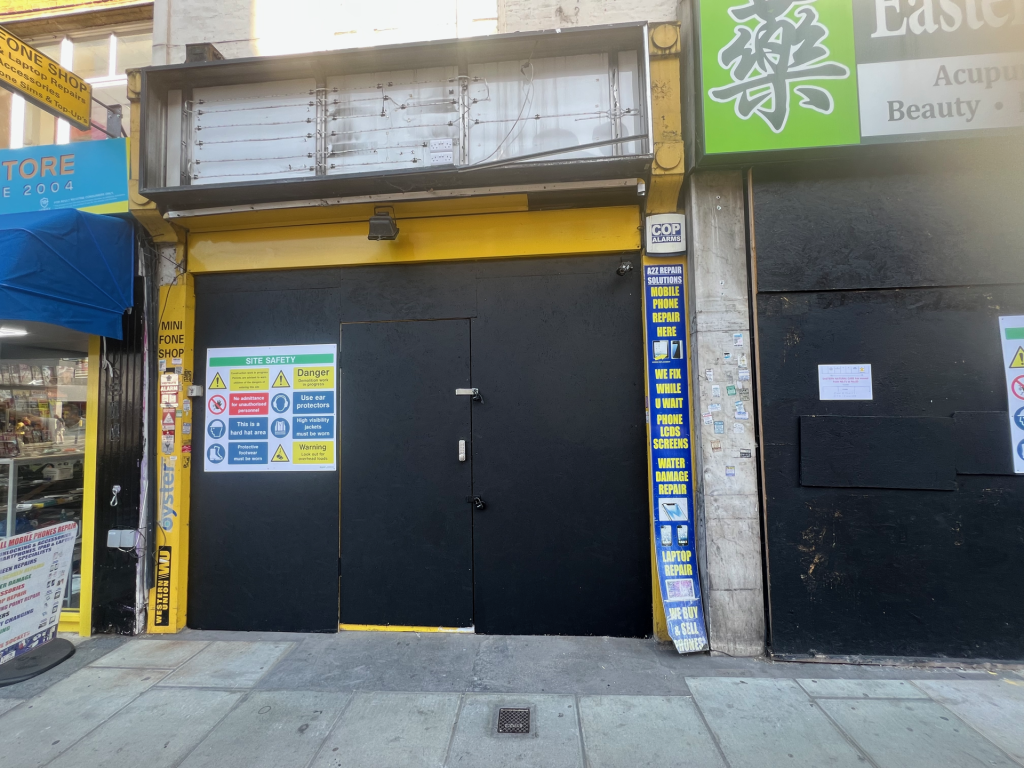 Closed storefront with a black metal door under a yellow frame; safety posters on the wall and a blue vertical ad for mobile phone repair beside it.