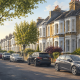Residential street lined with brick townhouses, trees, and parked cars on both sides on a sunny day.