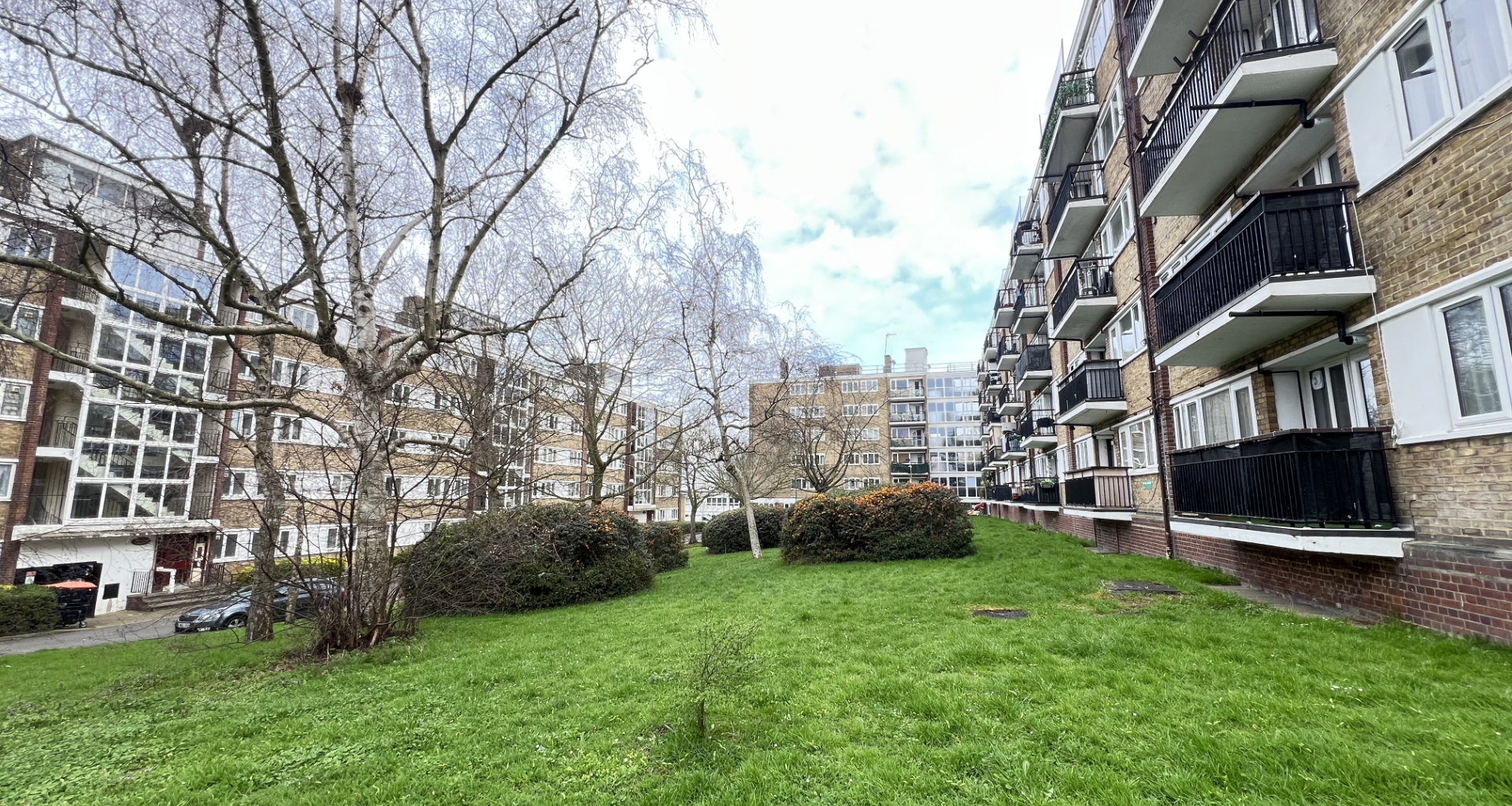 Urban residential courtyard: green lawn, bare trees, and multi-story brick apartment buildings with balconies on the right side.