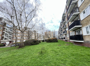 Urban residential courtyard: green lawn, bare trees, and multi-story brick apartment buildings with balconies on the right side.