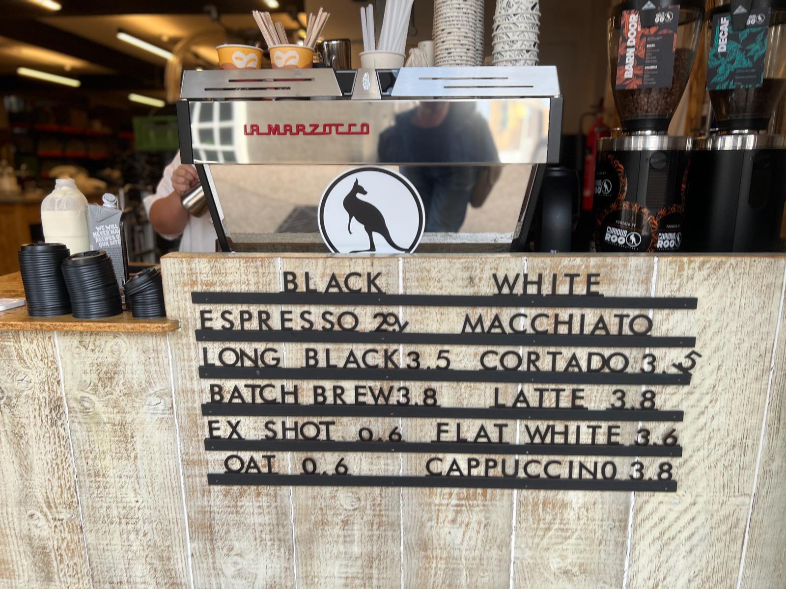 Letter board menu on a wooden counter listing espresso, long black, flat white at 3.6 and other drinks