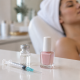 Close-up of a clear vial, syringe and pale pink nail polish on a white beauty-salon counter, with a woman reclining in a treatment chair softly out of focus in the background