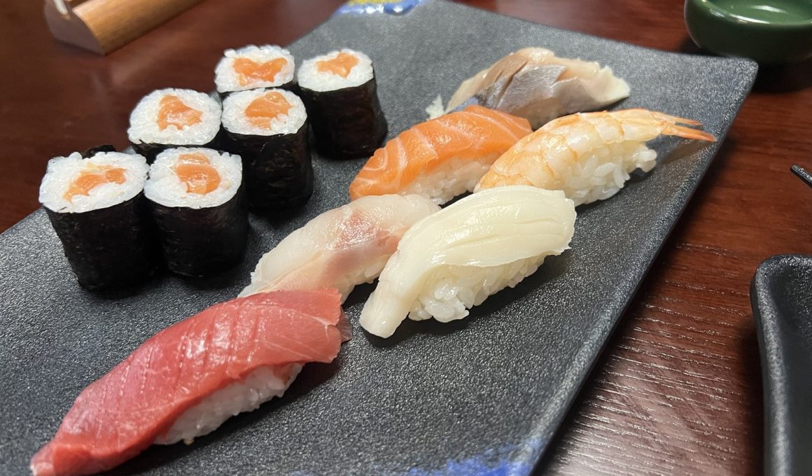 Six pieces of nigiri, including tuna, salmon and prawn, beside six salmon maki on a dark plate with a blue and gold glaze