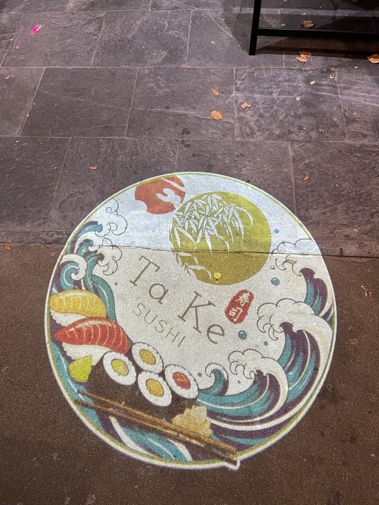 A round Ta Ke Sushi logo with waves, sushi and chopsticks, projected onto paving stones at night